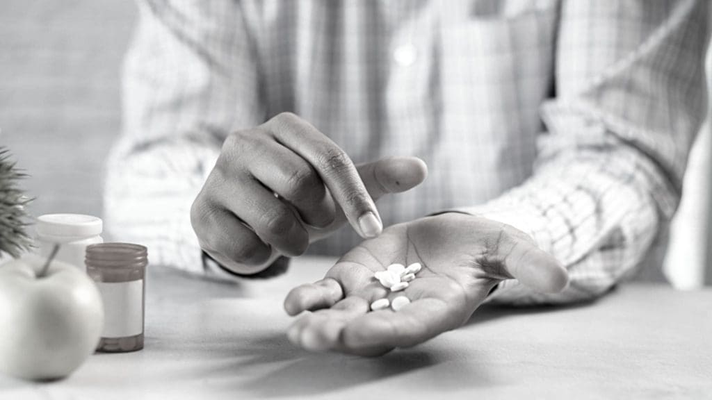 A pharmacist in the foreground with several pills in his palm and more bottles in the background.