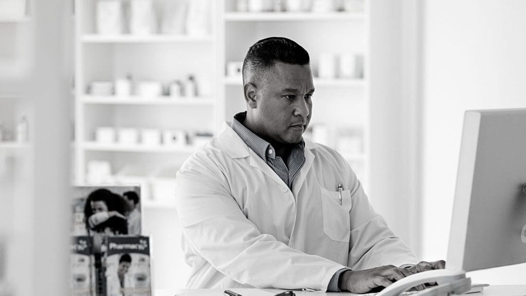 a male pharmacists working on a computer with drugs in background