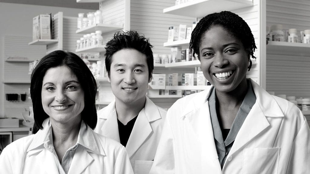 three pharmacists smiling infront of drugs in shelf on background
